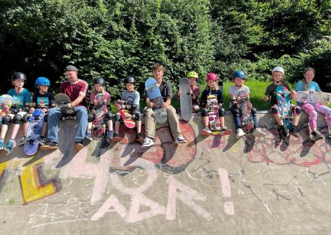 Eine Gruppe von Kids sitzt mit zwei Übungsleitungen, und jeweils einem Skateboard in der Hand, auf einer Rampe im Skatepark.