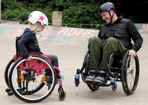 Ein Kind und ein erwachsener Mann sitzen sich in einem Rollstuhl, auf der Skateanlage Südstraße, zusammen. Der Erwachsene Mann zeigt dem Mann einen Skate-Trick im Rollstuhl.