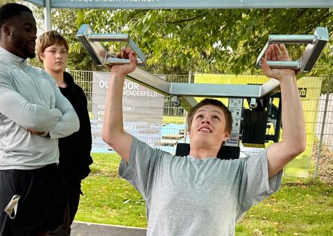 Ein Jugendlicher sitzt auf einem klassischen Fitnessgerät und führt die Schulterpresse aus. Zwei andere männliche Personen schauen ihm zu. 